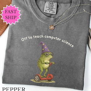 Può includere: Una maglietta grigia Comfort Colors con la scritta "Off to teach computer science". La maglietta presenta un'illustrazione stravagante di una rana con cappello da mago e occhiali, su uno scooter rosso. È visibile anche una collana d'argento con un ciondolo.