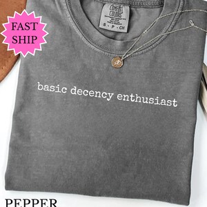 Può includere: Una maglietta grigia con la scritta "basic decency enthusiast" stampata in bianco. Una collana d'argento con un piccolo ciondolo è appoggiata sulla maglietta. La maglietta è piegata e ha un'etichetta "Comfort Colors".