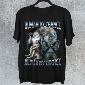 May include: Black t-shirt with a graphic design featuring three wolf-like figures, a full moon, and lightning bolts. The text "HUMAN BY CHANCE" and "ALPHA BY CHOICE" are displayed in a stylized font.