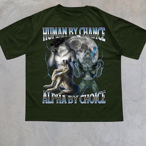 May include: Military green t-shirt with a graphic featuring three wolf-like figures in front of a full moon. The text "HUMAN BY CHANCE" and "ALPHA BY CHOICE" are displayed in white and blue.