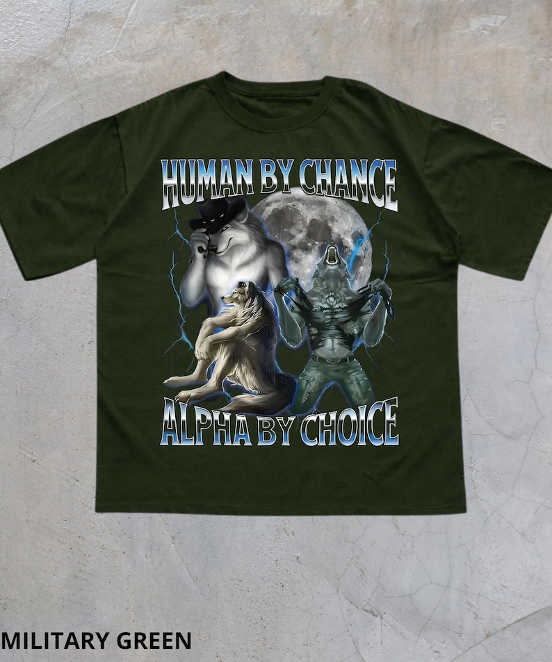 May include: Military green t-shirt with a graphic featuring three wolf-like figures in front of a full moon. The text "HUMAN BY CHANCE" and "ALPHA BY CHOICE" are displayed in white and blue.