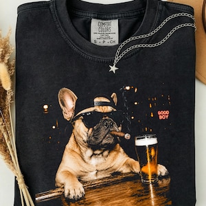 Peut inclure: Un t-shirt noir à col rond avec un imprimé représentant un bouledogue français portant une casquette et des lunettes de soleil, fumant un cigare et posant ses pattes sur un bar à côté d'une bière. Le texte GOOD BOY est écrit en rouge.