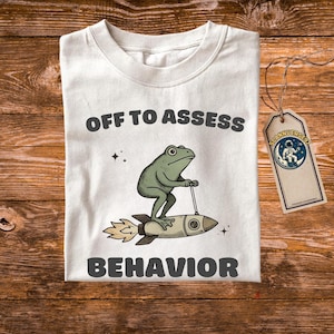 Könnte beinhalten: Weißes T-Shirt mit dem Text "OFF TO ASSESS BEHAVIOR". Das Shirt zeigt einen Cartoon-Frosch, der ein Raumschiff reitet. Das Shirt hat ein Etikett mit der Aufschrift "SCANNIVERSES97".