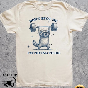 May include: Cream-colored t-shirt featuring a raccoon lifting a barbell with the text "DON'T SPOT ME I'M TRYING TO DIE" in navy blue. The design includes stars and a distressed aesthetic. The shirt is made of a soft, natural fabric.