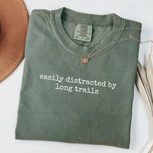 Può includere: Una maglietta verde muschio con la scritta bianca "easily distracted by long trails". La maglietta è piegata e ha una piccola etichetta Comfort Colors. Una collana d'oro con un ciondolo è appoggiata sulla maglietta. Un cappello marrone è parzialmente visibile.