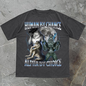 May include: Charcoal-colored t-shirt with a graphic design featuring a wolf wearing a hat, a wolf sitting down, and a creature in front of a full moon. The text "HUMAN BY CHANCE" and "ALPHA BY CHOICE" are displayed in blue.