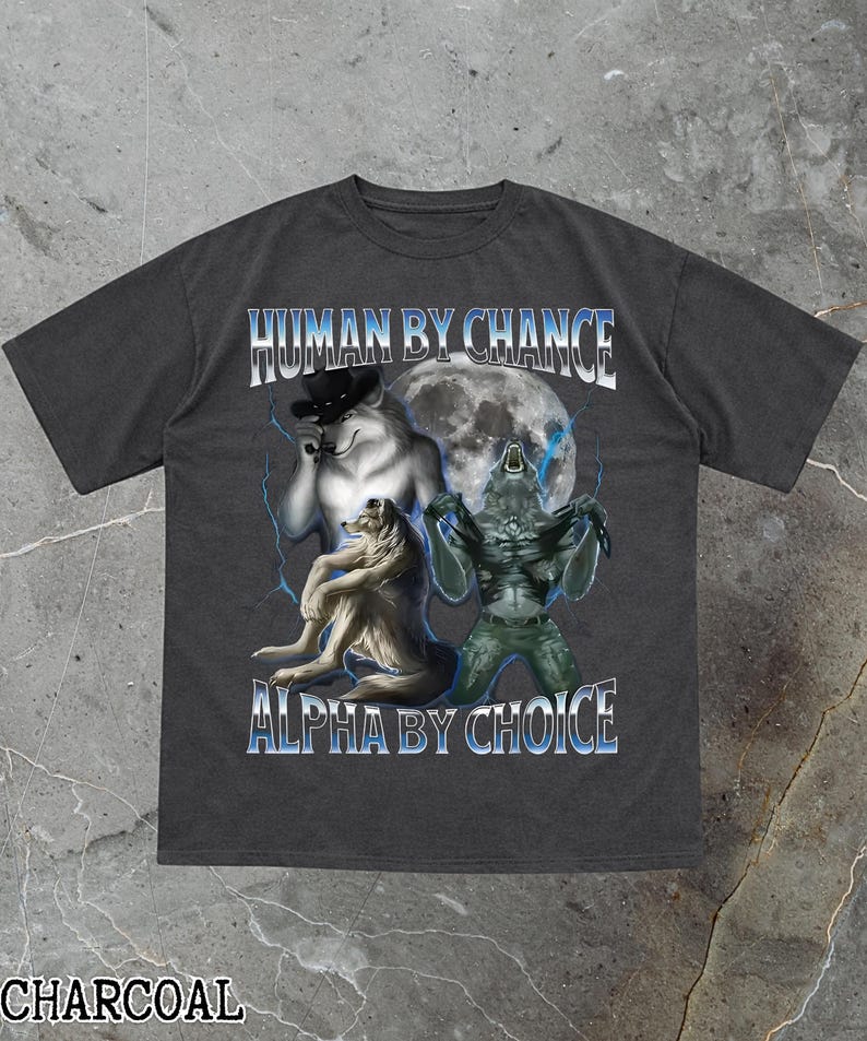 May include: Charcoal-colored t-shirt with a graphic design featuring a wolf wearing a hat, a wolf sitting down, and a creature in front of a full moon. The text "HUMAN BY CHANCE" and "ALPHA BY CHOICE" are displayed in blue.