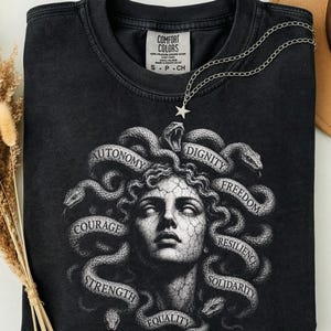 Peut inclure: Un t-shirt noir avec un motif représentant un visage à la texture de pierre entouré de serpents. Le design inclut les mots Autonomy, Dignity, Freedom, Resilience, Solidarity, Equality, Strength et Courage. Un collier étoile argenté repose sur le col.