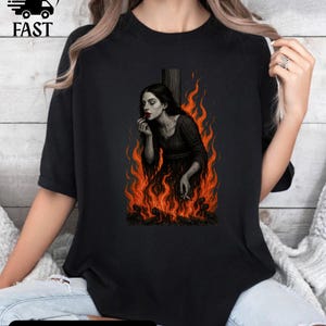 Op de afbeelding: Zwart T-shirt met een afbeelding van een vrouw in vlammen, lippenstift aanbrengend. De afbeelding heeft een vintage, gotische uitstraling. De tekst "COLOR: BLACK" staat onderaan en "FAST" linksboven.