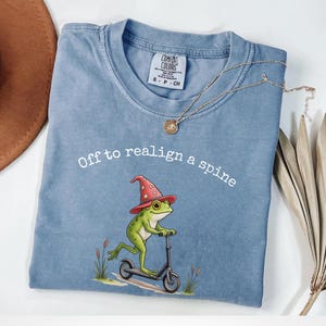 Puede incluir: Una camiseta azul Comfort Colors con el texto "Off to realign a spine". La camiseta presenta una rana de dibujos animados con un sombrero de mago y montada en un patinete. Un collar de oro está drapeado sobre la camiseta. La palabra "BLU JEAN" está en la parte inferior.
