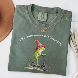 Op de afbeelding: Mosgroen t-shirt met een grillige kikker-afbeelding die een tovenaars hoed draagt en op een scooter rijdt. De tekst "Off to improve someone's flexibility" staat boven de kikker. Het shirt is van het merk Comfort Colors.