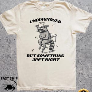 May include: Cream-coloured t-shirt with a black and white illustration of a raccoon wearing a cowboy hat and sitting on a toilet. The text reads "UNDIAGNOSED BUT SOMETHING AIN'T RIGHT".