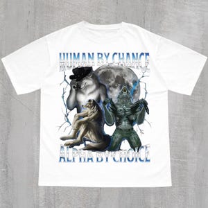 May include: White t-shirt featuring a graphic design with the text "HUMAN BY CHANCE" and "ALPHA BY CHOICE" in blue. The design includes illustrations of a wolf wearing a hat, a wolf, and a creature in front of a full moon.