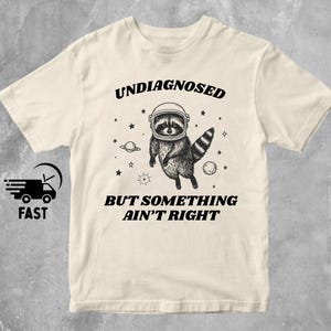 May include: Cream-coloured t-shirt with a raccoon in an astronaut helmet, surrounded by stars and planets. The text "UNDIAGNOSED BUT SOMETHING AIN'T RIGHT" is printed above and below the raccoon illustration. The shirt also has the word "NATURAL" printed at the bottom.