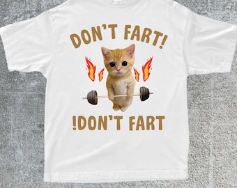 Pode incluir: Camiseta branca com um gráfico engraçado. O design apresenta um gato de desenho animado levantando uma barra, chamas e o texto "DON'T FART!" acima e abaixo do gato. A camisa é um estilo casual unissex.