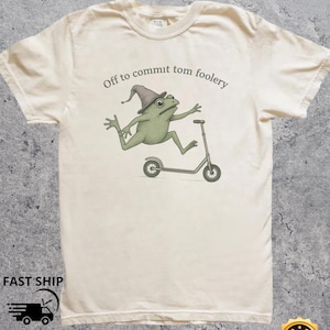 Könnte beinhalten: Cremefarbenes T-Shirt mit einem grünen Frosch, der einen Zaubererhut trägt und auf einem Roller fährt. Der Text "Off to commit tom foolery" ist über dem Frosch aufgedruckt. Das Shirt hat einen Rundhalsausschnitt und kurze Ärmel.
