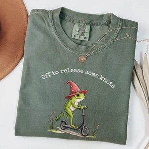 Könnte beinhalten: Moosgrünes T-Shirt mit einer skurrilen Froschgrafik. Der Frosch, der einen roten Zaubererhut trägt, fährt einen schwarzen Roller. Der Text "Off to release some knots" ist darüber. Marke Comfort Colors.