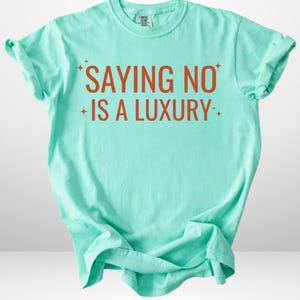 Puede incluir: Camiseta turquesa clara con el texto naranja "SAYING NO IS A LUXURY". La camiseta tiene mangas enrolladas y cuello redondo. Las palabras están acentuadas con gráficos de estrellas.
