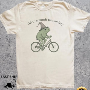Könnte beinhalten: Cremefarbenes T-Shirt mit einer grünen Froschfigur auf einem Fahrrad, die einen Zaubererhut trägt. Der Text "Off to commit tom foolery" ist über dem Frosch aufgedruckt. Das Shirt hat einen Rundhalsausschnitt und kurze Ärmel.