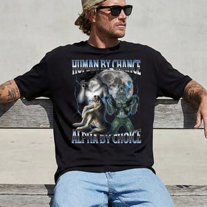 May include: Black t-shirt with the text "HUMAN BY CHANCE ALPHA BY CHOICE" in blue and white. The graphic features a wolf, a creature, and a full moon with lightning bolts. The shirt is worn by a person.