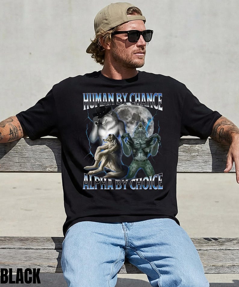 May include: Black t-shirt with the text "HUMAN BY CHANCE ALPHA BY CHOICE" in blue and white. The graphic features a wolf, a creature, and a full moon with lightning bolts. The shirt is worn by a person.