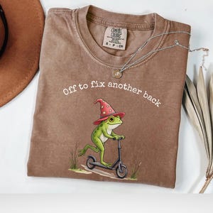Op de afbeelding: Espresso-kleurig T-shirt met een groene kikker met een tovenaars hoed op een scooter. De tekst "Off to fix another back" staat boven de kikker. Het shirt heeft een Comfort Colors label.