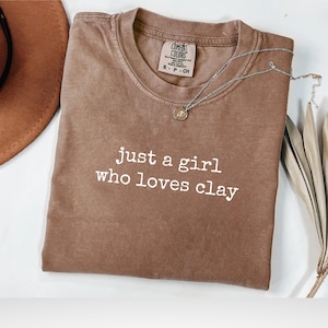 Può includere: T-shirt color espresso con la scritta bianca "just a girl who loves clay". Una collana d'argento con un ciondolo è appoggiata sulla maglietta. Un cappello marrone e foglie secche sono visibili sullo sfondo.