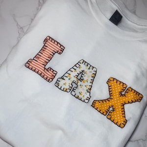 Op de afbeelding: Wit t-shirt met de letters "LAX" op de voorkant. De "L" is oranje met witte strepen, de "A" is wit met een geel bloemenpatroon en de "X" is oranje met witte stippen.