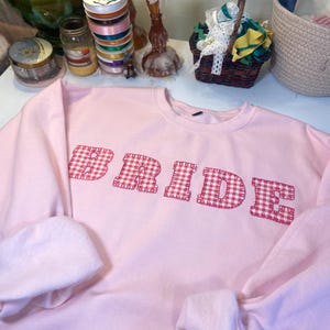 Może przedstawiać: Jasnoróżowa bluza z napisem "BRIDE" wyszytym na piersi w czerwono-białą kratę. Bluza ma długie rękawy i okrągły dekolt. Inne przedmioty na zdjęciu to wstążki, świece i kosz.