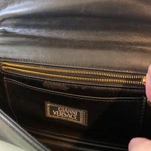May include: Black leather clutch with a gold zipper and the "Gianni Versace" logo. The interior features a pocket and a gold zipper. The clutch is made of a smooth, shiny leather.