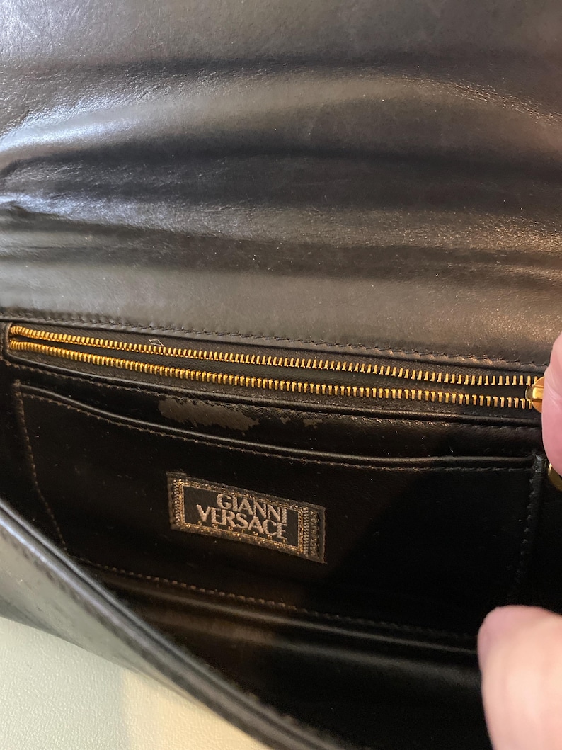 May include: Black leather clutch with a gold zipper and the "Gianni Versace" logo. The interior features a pocket and a gold zipper. The clutch is made of a smooth, shiny leather.