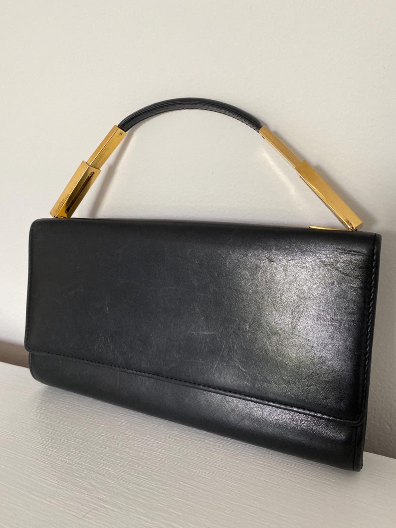 A black leather clutch with a gold metal handle. The clutch has a flap closure and a smooth, textured surface.