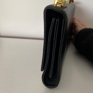 May include: Black leather briefcase with a gold-toned clasp. The briefcase has a rectangular shape with a structured design. The clasp is rectangular and has a gold finish. The briefcase is closed.