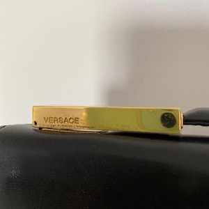 May include: A gold-toned rectangular clasp with the word "Versace" engraved on it. The clasp is attached to a black leather surface. The image is a close-up shot, highlighting the details of the clasp and the texture of the leather.
