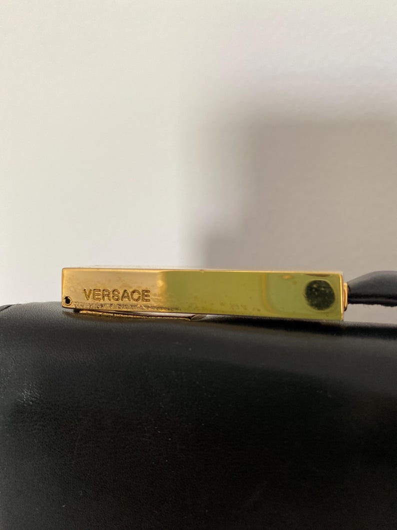 May include: A gold-toned rectangular clasp with the word "Versace" engraved on it. The clasp is attached to a black leather surface. The image is a close-up shot, highlighting the details of the clasp and the texture of the leather.