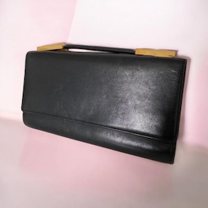 May include: A black leather clutch with a rectangular shape. It features a handle and gold-colored accents on the top. The clutch has a flap closure and a smooth, textured surface.