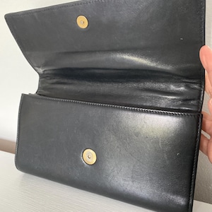 May include: Black leather clutch with a gold-tone clasp. The clutch is open, revealing the interior. The leather appears smooth and has a slight sheen. The clutch is rectangular and has a simple, classic design.