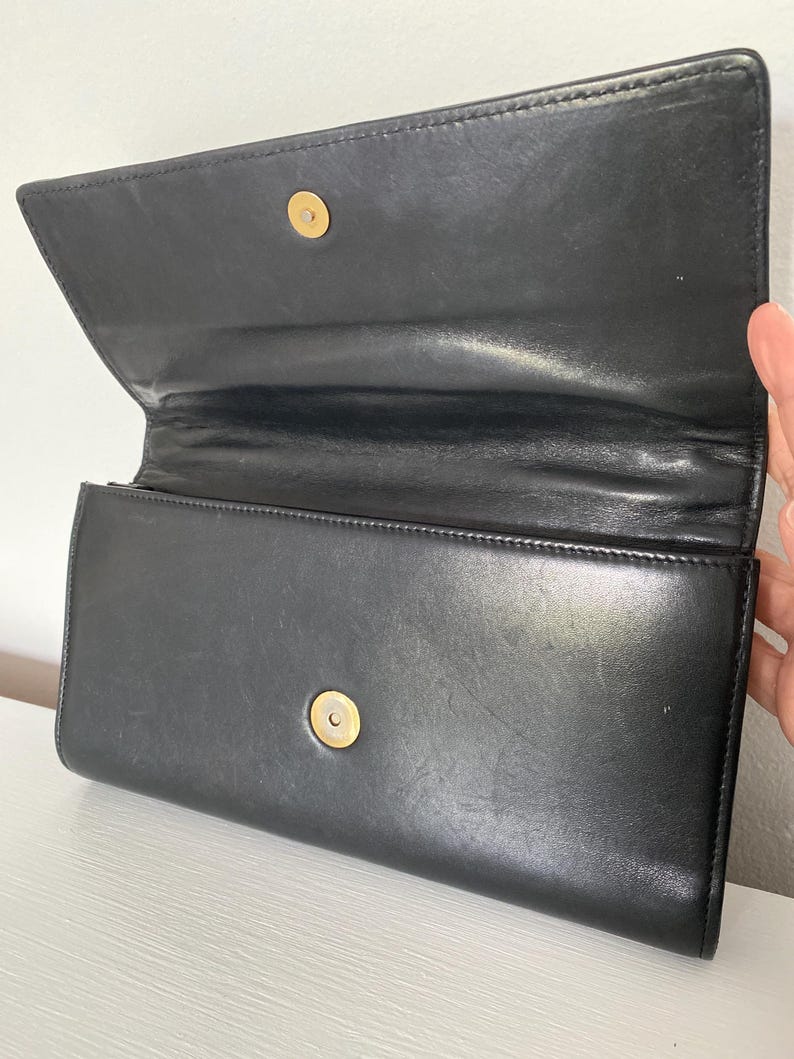May include: Black leather clutch with a gold-tone clasp. The clutch is open, revealing the interior. The leather appears smooth and has a slight sheen. The clutch is rectangular and has a simple, classic design.