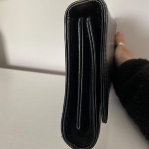May include: Black leather clutch with a gold-colored clasp. The clutch is rectangular and has a folded design. The image shows the side view of the clutch.
