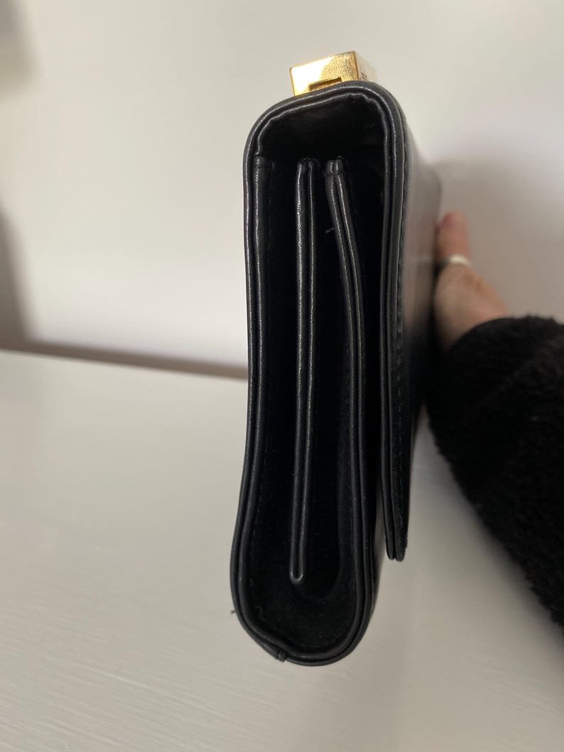 May include: Black leather clutch with a gold-colored clasp. The clutch is rectangular and has a folded design. The image shows the side view of the clutch.