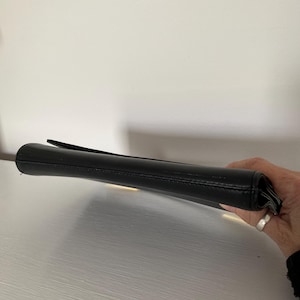 May include: A black clutch purse with a flap closure. The purse is made of a smooth material and appears to be empty. The clutch is held by a person's hand.