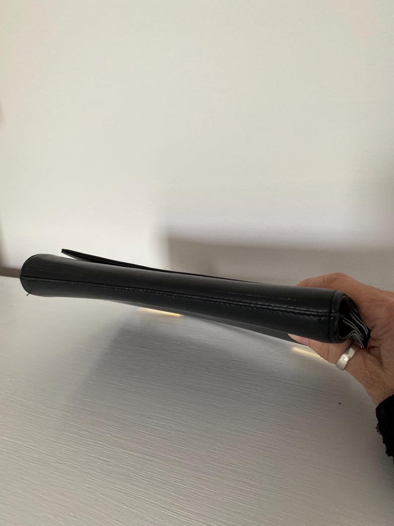 May include: A black clutch purse with a flap closure. The purse is made of a smooth material and appears to be empty. The clutch is held by a person's hand.