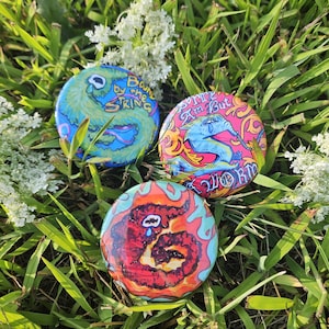 May include: Three round, colorful buttons with unique designs and text. One button features a blue and green design with the text "Bound by the String." Another has a pink background with a blue elephant and the text "Sorry I'm But Worm." The third button has a red worm design.
