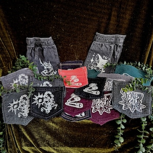 May include: A collection of denim pockets in various colors including black, gray, red, teal, and maroon, featuring white screen-printed graphics of safety pins, spiked chains, and the text "PUNK KAT" alongside two pairs of gray denim pants.