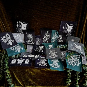 May include: A collection of rectangular fabric patches in black, dark gray, and teal, each featuring white screen-printed illustrations of cats, unicorns, daggers, and text including Beck's Trinket Emporium and Pink Kat, arranged in a cluster.