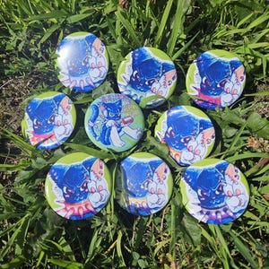 May include: A collection of round, glossy buttons with cartoon illustrations. The buttons feature a blue and white creature with an angry expression. One button has the text "Trans Rights Are Human Rights" on a green background.