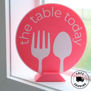 May include: A pink circular sign with the text "the table today" in white. The sign features white fork and spoon silhouettes. The sign is on a pink base and is placed on a windowsill. A "Free Shipping" sticker is in the bottom right corner.