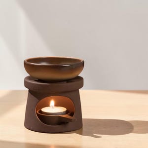 May include: A brown ceramic essential oil diffuser with a small bowl on top and a tea light candle burning inside. The diffuser is sitting on a light brown wooden surface.