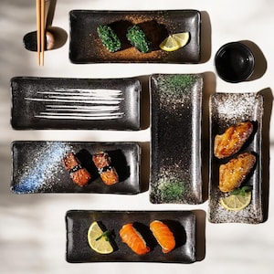 Puede incluir: Conjunto de platos rectangulares negros de cerámica con diversos alimentos, como sushi, pollo frito y rodajas de limón. Algunos platos tienen diseños moteados blancos o verdes. También hay palillos y un cuenco negro pequeño.