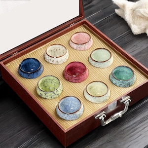 Puede incluir: Un juego de nueve pequeñas tazas de té de cerámica en varios colores, incluyendo azul, verde, rosa y blanco, dispuestas en una caja de madera con un interior dorado. Las tazas tienen un aspecto texturizado y moteado.